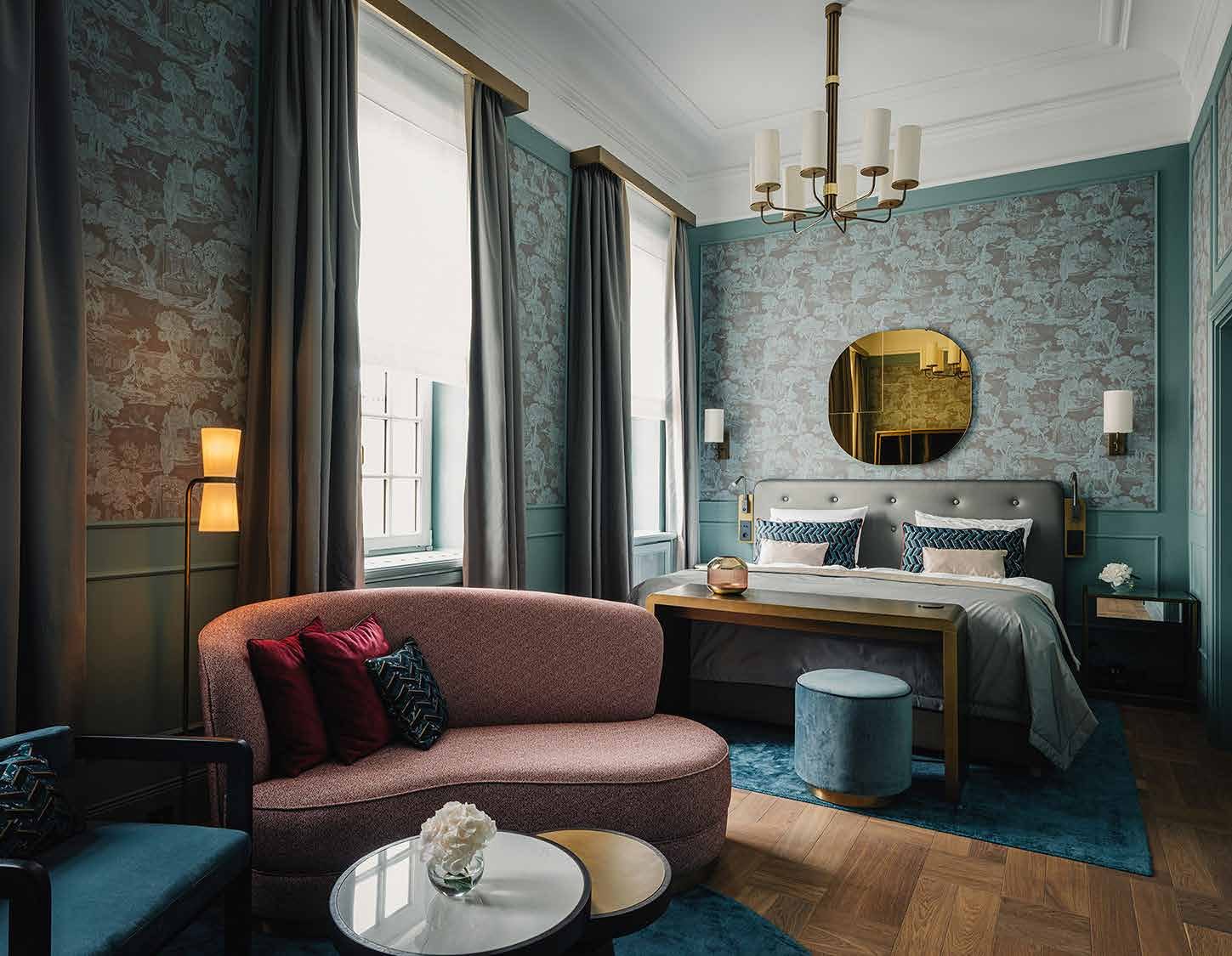Zimmer im Designtyp II: türkisgrüne Wände mit Bildtapete, rosa Sofa, runder Messingspiegel über dem Bett und blauer Teppich