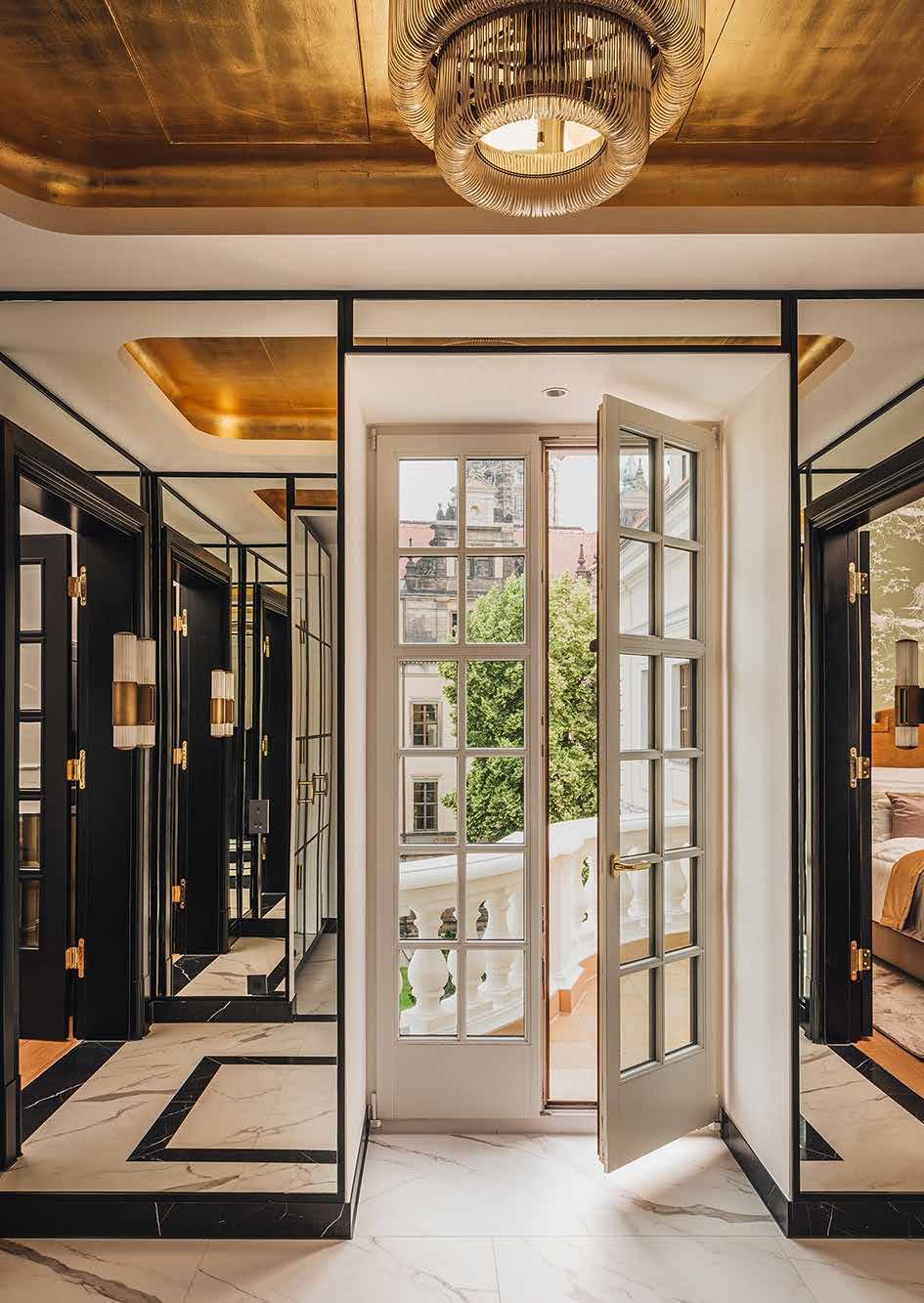 Raum im sanierten Taschenbergpalais: goldene Decke mit Glasleuchter, schwarz gerahmte Spiegel- und Glastüren, Marmorboden mit schwarzen Intarsien und eine geöffnete weiße Sprossentür zum Balkon mit Blick auf die Dresdner Altstadt