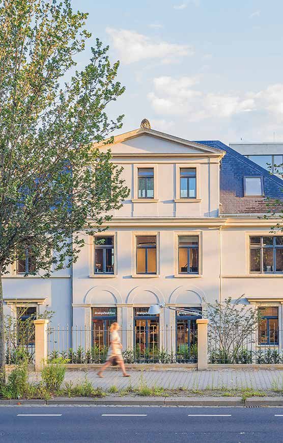 Straßenansicht der sanierten Behr’schen Villa in Dresden: hell verputzte Fassade mit Dreiecksgiebel und Rundbogenfenstern im Erdgeschoss, davor Zaun, Gehweg und eine vorbeigehende Passantin
